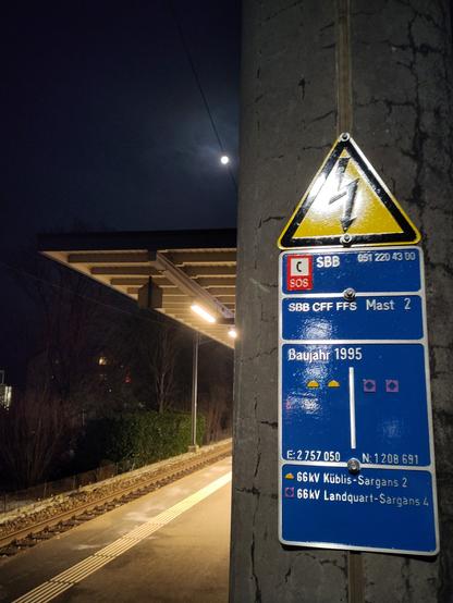Foto. Bahnhof, nachts, mit Schildern einer Hochspannungsleitung.