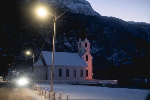 A norwegian white chapel by the side of the road, in a valley in the middle of the mountains, snow in the ground.