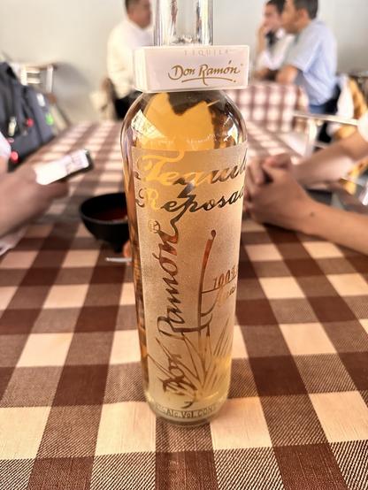 A bottle of Don Ramón Reposado tequila is placed on a checkered tablecloth in a pozolería. In the background, there are blurred figures of people engaged in conversation.