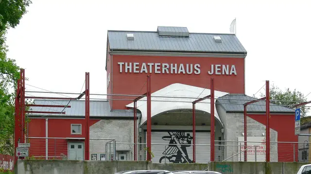 The image depicts a building with a sign that reads "THEATERHAUS JENA." It appears to be a theater or performance venue, featuring a distinctive architectural design with a red exterior. The structure has a modern appearance, with a roof that includes noticeable overhangs and a parking area in front. There are also some artistic elements visible on the exterior.

Image Credits: Wikimedia / Photo: Andreas Praefcke / CC BY 3.0