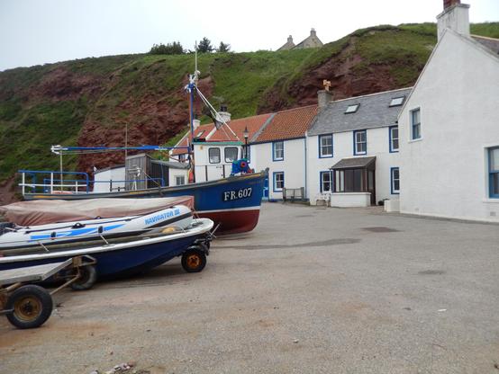 A blue and white fishing boat labeled "FR 607" is docked in front of several white houses with blue window frames. A smaller blue and white inflatable boat named "Navigator II" sits on a trailer in front of the larger vessel. A grassy hill with a reddish cliff face is visible in the background.