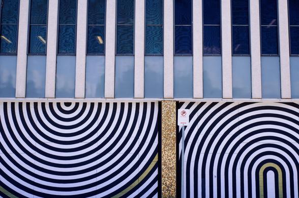 Photograph of the side of a building.  The top floor has a an alternating pattern of vertical windows and concrete columns.  The bottom floor is painted with circular patters of black and white stripes.  A No Parking sign is in the middle of the frame, in front of the ground floor.