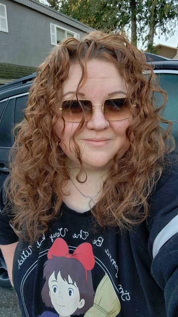 Me, a very pale white woman with red hair is smiling at the camera. I'm wearing sunglasses and a Kiki's Delivery Service sweater. My curly hair is looking awesome, lots of volume, some definition.