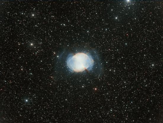 Sur un fond de ciel tapissé d'étoiles, la nébuleuse planétaire Messier 27, surnommée Dumbbell, Haltère, Battant de cloche, Trognon de pomme, Sablier, Diabolo… De part et d'autre de la partie centrale un peu surexposée de forme ovale et de couleur blanche, de faibles extensions bleues apparaissent. Un faible fuseau rouge semble transpercer le cœur de la nébuleuse.