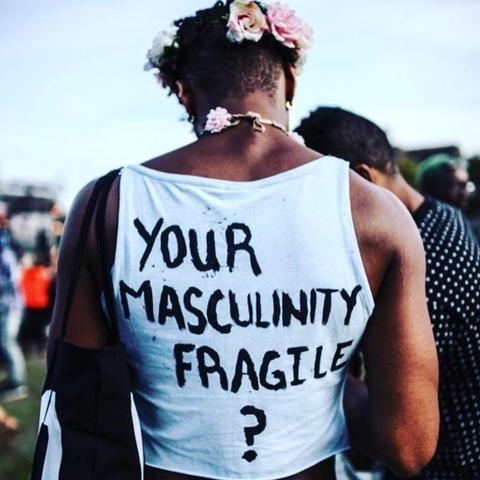 Photo of a man's back. He is muscular, has flowers in his hair, and has "Your masculinity fragile?" written on his shirt.