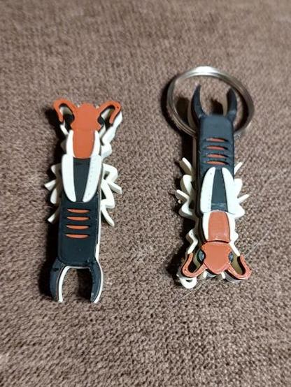 Two 3d printed Lined Earwig designs. The have a reddish head and the thorax is the same reddish color with tan stripes along the outside edges. The abdomen is black with black pincers and reddish stripes. The wing covers are black with tan stripes along the outer edges. The legs are tan. The left earwig is facing the top of the picture. The right earwig is facing the bottom of the picture with a 1-inch (2.5cm) spilt ring through the abdomen.