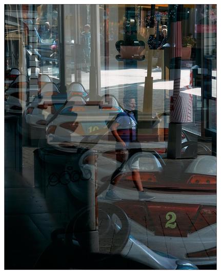 This image is a visually complex, multi-layered scene, possibly a reflection or taken through glass, showing various elements superimposed over each other. You can see a coin-operated children's ride shaped like cars, with prominent numbers "10" and "2." There's a reflection of a street scene that includes pedestrians, a pole, and buildings with shopfronts. The overlay of images creates an intriguing, somewhat disorienting effect, merging the interior and exterior environments.