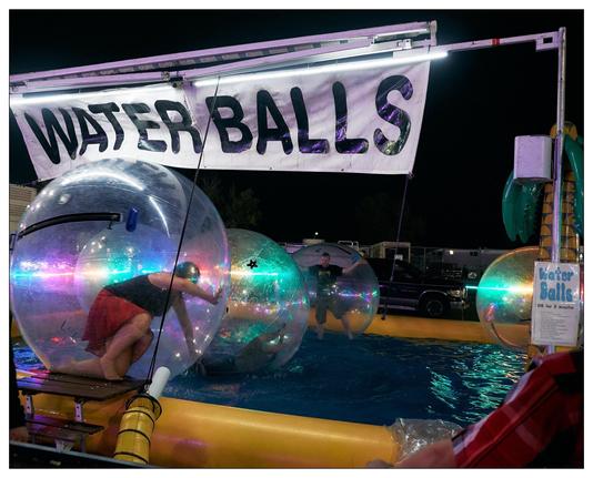 This image captures a fun and colorful scene at what appears to be a fair or amusement park. It shows an activity area titled "Water Balls," where people can go inside large inflatable balls that float on water. The image features two individuals inside these transparent orbs, floating in a small pool outlined with yellow barriers. One person is actively moving inside the ball, attempting to walk or roll it across the water, while the other appears to be standing still, possibly adjusting to the space or taking a moment to enjoy the activity. The area is illuminated by ambient and colorful lighting, which adds a lively and vibrant atmosphere to the scene.