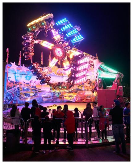 This image captures a lively scene at a carnival or amusement park at night. Central to the photo is a large, brightly lit amusement ride featuring multiple arms with passenger cars that spin and rotate, creating a dynamic and thrilling experience. The ride is adorned with flashing lights and neon signs that add to its eye-catching appearance.

In the foreground, spectators, possibly families or groups of friends, are standing behind a barrier, observing the ride. Some individuals are directly watching the ride, while others appear to be interacting with each other. The setting suggests a joyful and exciting atmosphere typical of such events, emphasizing entertainment and community engagement. The backdrop of the ride features vibrant artwork, contributing to the colorful and energetic environment.