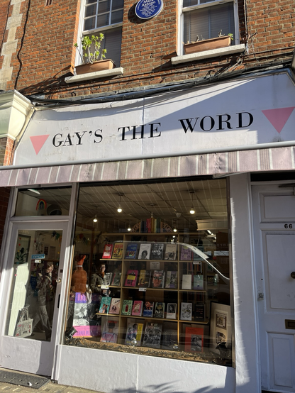A bookstore window, filled with books on display. Above the window is a marquee sign with black letters on a white background, reading GAY’S THE WORD in capital letters. The name is flanked on each side by an upside down pink triangle.