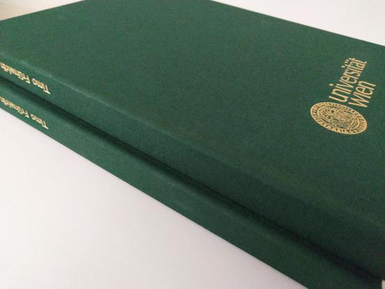 Two dark-green hardcover books are stacked on top of each other.  The top book has "Universität Wien" and a gold seal embossed on the cover.
Credits to @altbot.