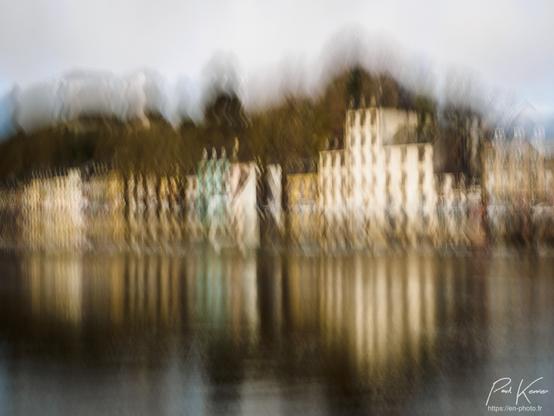 Photo en couleur, présentant les maisons d'un quai et leur reflet dans un canal sous un ciel couvert. La photo avec une certaine longueur d'ouverture de l'obturateur a été captée avec un mouvement volontaire vertical afin de générer du flou de bougé et allonger la hauteur des bâtiments, la plupart de ceux-ci étant peints en blanc ou beige clair à l'exception de l'un d'entre eux peint en bleu .