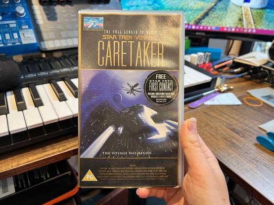 A photo of a VHS copy of the debut two-part episode of Star Trek: Voyager, Caretaker. The cover is mostly black with a gold print border. The Paramount logo is at the top left, there’s a Star Trek 30 Years logo at the top right and at the top it says ‘THE FULL LENGTH TV MOVIE’ in a white narrow font, and under that ‘STAR TREK : VOYAGER’ in gold in the show’s logo font. Under them both is ‘CARETAKER’ in a large, narrow gold font.  There is an image of the USS Voyager flying towards the Caretaker array which covers most of the front, with ‘THE VOYAGE HAS BEGUN…’ at the bottom of the image. The PG rating is at the bottom left and the credits are at the bottom in a small, narrow gold font.