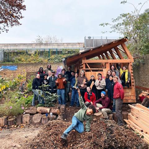 A group of local people stand outside the partly completed barn they are building as a community space. A child is jumping on the pile of earth at the front of the group.