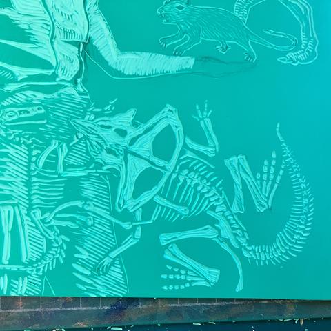 Detail of my green vinyl Lino block being carved showing a protoceratops fossil next to a standing person and part of a velociraptor fossil. The woman’s torso and one extended arm is visible. Her hand remains uncarved but above it is a rodent.