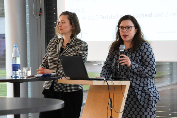 Ministerialdirektorin Leonie Dirks spricht in ein Mikrofon. Daneben steht Referatsleiterin Dr. Layla Distler.