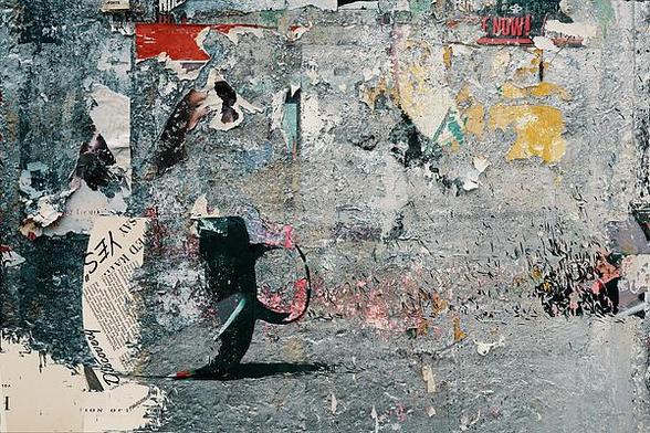 A digital collage composed of grungy urban walls with peeling posters and such, including a red-on-black piece stating "NOW!"  and some newsprint. The focal element is a coffee mug with similar texture.
