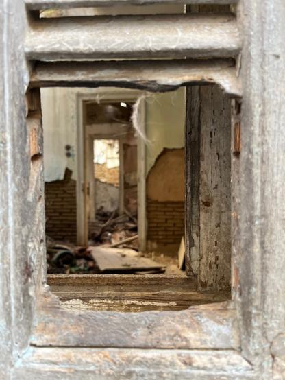 Through the frame of a broken door panel, the out of focus interior of an abandoned house. A room with a floor strewn with beams and debris, the far wall showing brick and mortar below, plaster above, including a bit of decorative paintwork to the left of an interior door aligned with our viewpoint and a further door to the courtyard outside. All in shades of mud and nicotine brown, with a bit of yellowing cream where the plaster remains. It’s the three openings that make it work, the broken outer door, the fallen door between two rooms straddled by its doorframe, the glimpse of daylight through the far doorway.