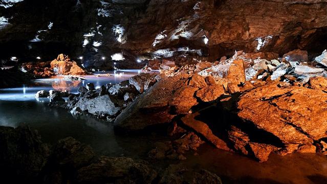 Photo in a dark cave, showing the rough ceiling, walls and big rocks on the foreground. There's orange light illuminating the closest rocks while smaller white spotlights shine towards the lower parts in the back and shining onto a water surface on the left side. The whole scene creates a beautiful atmosphere and depth to an otherwise dark cavern, almost magical.