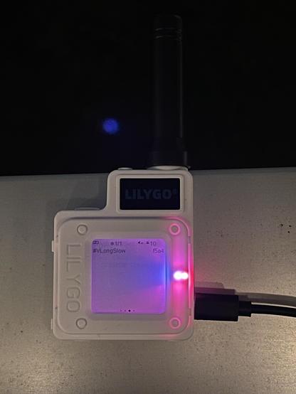 A small white electronic device (Lilygo T-Echo Meshtastic) with a screen displaying data, two LED indicator lights (one red and one blue), and an antenna. It is connected to a cable on a metallic surface.