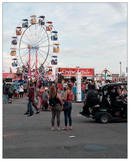 The image shows a lively carnival scene featuring a large Ferris wheel with colorful gondolas against a cloudy sky. In the foreground are people strolling and socializing in an area surrounded by various food stalls, including one named "Crepe Fair." There are two women in the center facing towards each other and talking; others are walking or gathering around. Additionally, several carnival rides and game booths can be seen in the background, creating a festive and vibrant atmosphere typical of a fairground.