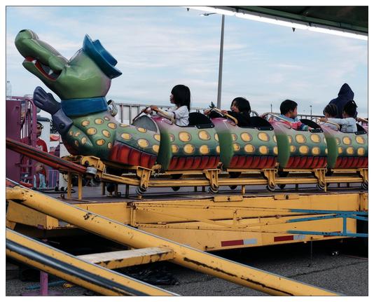 This image captures a colorful amusement park ride resembling an alligator. The structure includes a segmented body in yellow, green, and red with circular patterns, a smiling face, and a hat-like element on the head adding a whimsical touch. Children are seated in the compartments of the ride, secured by safety bars, and are seemingly enjoying their time as the ride operates on a track. The scene is set outdoors under an overcast sky, contributing to the mild lighting in the image. In the background, a supervising adult and some operational details of the ride, like stairs and control panels, are visible.