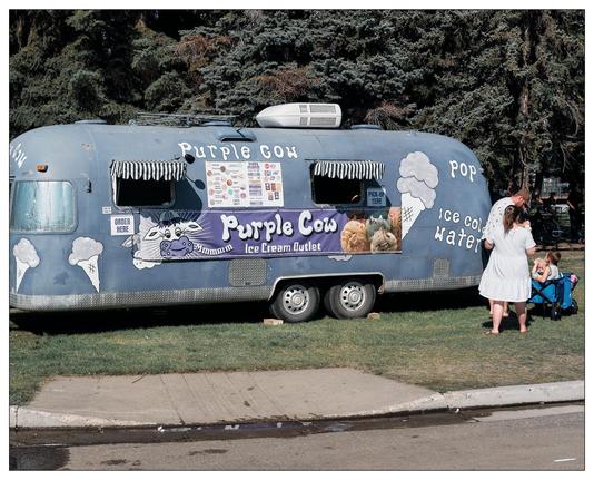 This is an image of a blue food truck called "Purple Cow Ice Cream Outlet" parked next to a grassy area with trees. The truck is decorated with images and text advertising various treats, such as ice cream, pop, and ice cold water. In front of the truck, there is a woman wearing a white dress standing with two children; one child is in a light-colored dress, and the other is seated in a stroller. The scene looks to be set in a sunny, outdoor area, indicating a casual, family-friendly atmosphere.