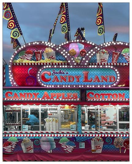 This image shows a brightly colored food concession stand named "Jackie's Candy Land." The stand displays various classic fair treats such as candy apples, cotton candy, popcorn, and snow cones. The decoration is vibrant and festive, with swirling patterns and bold, illuminated text, creating an enticing atmosphere commonly found in fairs or carnivals. There's also a worker visible inside the stand, likely preparing or selling the treats. The overall scene is lively and attractive, clearly designed to draw in customers at a fair or similar event.