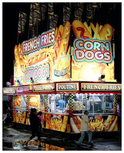 This image features a colorful and brightly lit food concession stand at a fair or carnival. The stand is adorned with vibrant graphics advertising a variety of popular snack foods such as French fries, hot dogs, corn dogs, chicken strips, nachos & cheese, and poutine. The imagery on the booth is quite eye-catching, featuring large representations of the foods offered, and decorative, flashy lights that enhance its appeal. In the foreground, there are a few people walking past, suggesting an outdoor setting, likely in the evening as the ground appears wet, possibly due to recent rain. The overall atmosphere conveyed is lively and festive, typical of a fairground setting.