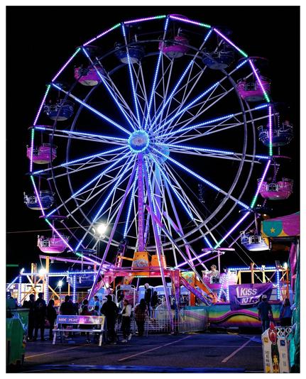 The image depicts a vibrant amusement park scene at night featuring a large Ferris wheel illuminated by colorful LED lights. The Ferris wheel has gondolas that are also lit, enhancing the festive and lively atmosphere. In the foreground and around the Ferris wheel, there are various amusement rides and booths, each adorned with bright lights and colorful decorations. People are visible enjoying the attractions, adding to the lively ambiance of a typical funfair or carnival setting. Signs for games and other entertainments, such as "Kiss 108" and other decorations, indicate a festive environment, likely involving music and various other activities typically found at fairs.