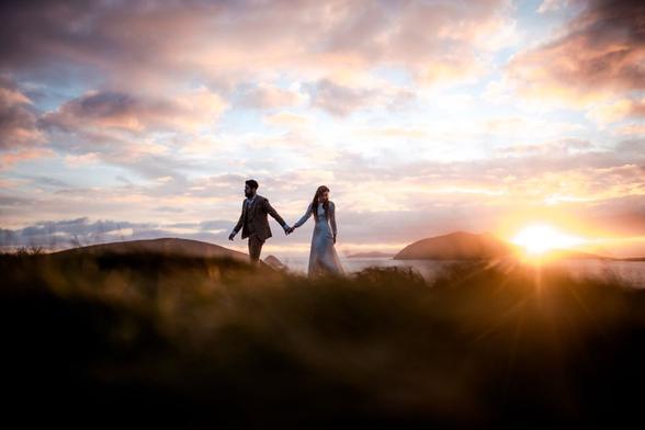 This image captures a couple holding hands, walking along a grassy cliff during sunset. The man is wearing a suit, and the woman is in a flowing dress. They appear calm and connected as they move forward together. In the background, the sun is setting behind distant hills, casting a warm, golden light over the landscape. The sky is painted with soft clouds in shades of blue, pink, and orange. The scene feels peaceful and intimate, surrounded by nature, with the ocean and rolling hills adding to the secluded atmosphere. This moment was captured by Hamburg wedding photographer Sturmsucht during an elopement on the Irish coast.