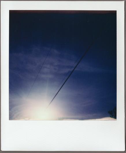 Polaroid looking up at a mostly clear, dark blue sky; three power lines run from the burst of the sun in the lower left and fade into the bluish black on the top right.