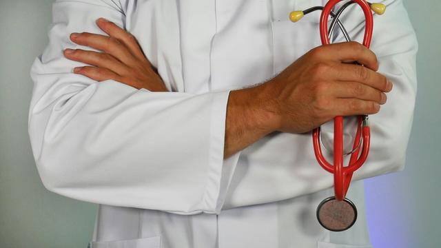 Zdjęcie lekarza w białym fartuchu, trzymającego ręce skrzyżowane na piersi. Kadr przedstawia tylko środkową część ciała lekarza i trzymane w lewej ręce słuchawki lekarskie.