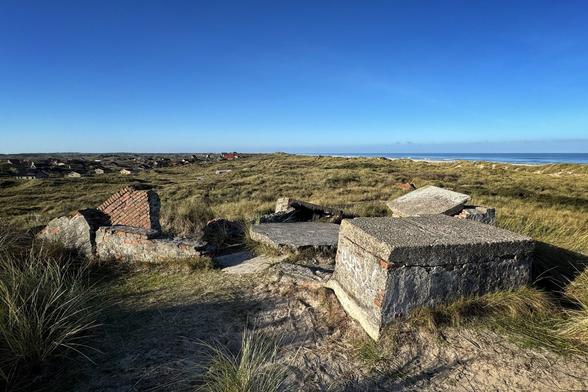 Partially collapsed concrete bunkers overgrown by dune sand and beach grass in the dunes near the beach of Midsland-Noord on the Dutch island of Terschelling. A holiday home settlement can be seen in the distance on the left, while a wide sandy beach stretches out behind the dunes in the sunshine and blue sky on the right.