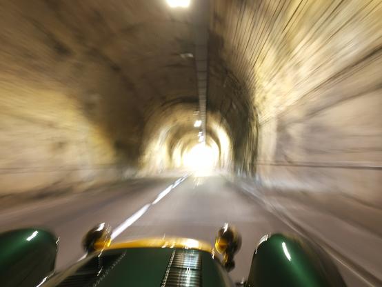 Photographie numérique couleur en format paysage.
Dans un tunnel arrondi, une voiture au capot allongé, avec un nez jaune et de grandes ailes vertes, roule vers la sortie qu'on voit au loin. Tout est flou : la voiture, les parois, la route.