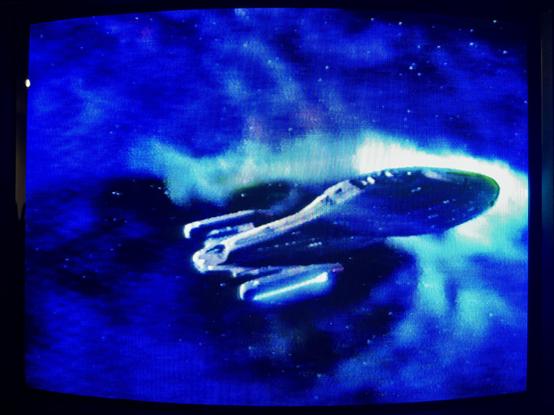 A photo of a CRT TV showing the opening titles of Star Trek: Voyager as played from the VHS cassette of the series’ first episode ‘Caretaker’. Here, the USS Voyager is flying though a nebula cloud, which is glowing as it comes into contact with the ship.