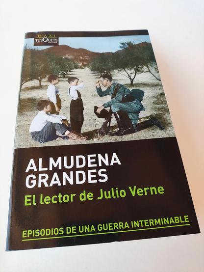 Portada dun libro en edición branda: "El lector de Julio Verne", de Almudena Grandes, un dos volumes da sua serie "Episodios de una guerra interminable". A cuberta é negra e nela hai unha imaxe dun garda civil nun olivar, cun xeonllo a terra, presignándose coa man dereita mentres sostén o tricornio na esquerda. Fronte a el, tes pequenos con camisas brancas e pantalóns longos, un deles sentado no chan abrazando as pernas, os outros en pé. Dos que están de pé, un deles está máis adiantado e imita ao adulto facendo o xesto da cruz sobre a súa fronte. Completa a cuberta o nome da autora en maiúsculas brancas, e o da obra e colección en verde.