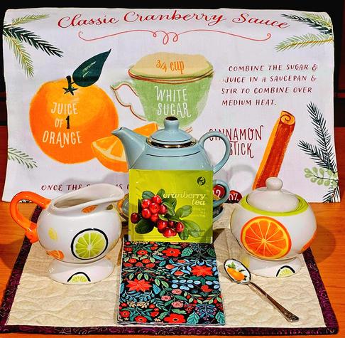 A tea tableau dedicated to cranberries and cranberry sauce. There is a tea towel printed with a classic recipe, creamer and sugar bowl with citrus motif for the zest in the recipe, a spoon with a painted orange, a napkin with cranberry colors. The tea is, of course, Cranberry black tea, from Adagio.