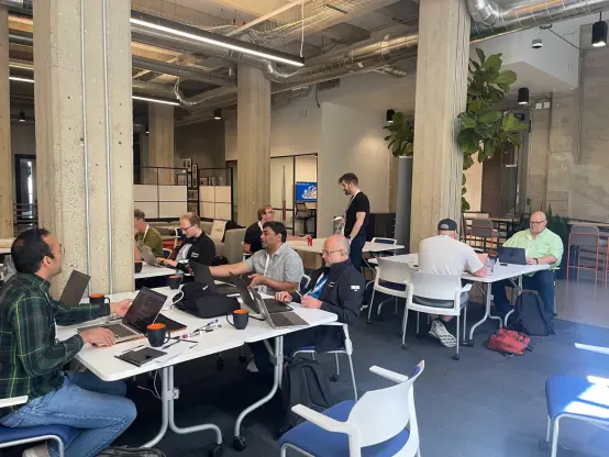 CommCon hackathon in progress with attendees sat at tables working on laptops and chatting