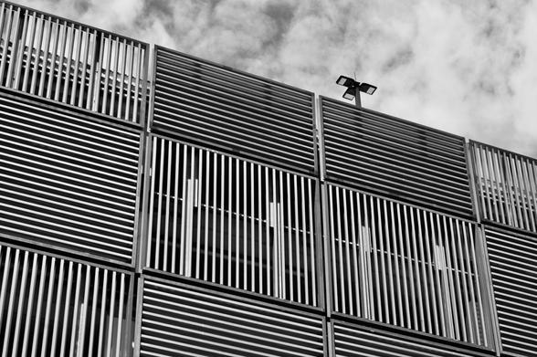 Black and white photograph looking up at a multi-storey car park.  The side of the carpark is made of square patches of steel strips. Each patch has strips running vertically or horizontally, making a chequerboard pattern on the side of the building.  A light pole is peeking above the top floor.