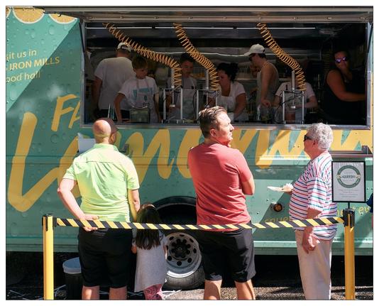 This image shows a scene at a food truck, which is decorated in a bright teal color with yellow accents. There are several customers standing in front of the truck. Two men and a woman are engaged in conversation; another man and a young child are also present, all of them appearing to be waiting or having just ordered food. Inside the food truck, multiple workers are visible, busy preparing orders. There are several coiled yellow air hoses attached to lemon presses.  The environment suggests a casual, sunny day, possibly at a public event or a market.