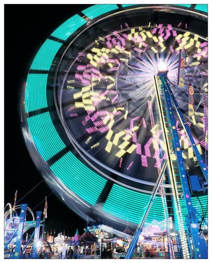 This image captures a vibrant and bustling amusement park at night, featuring a large Ferris wheel in motion. The Ferris wheel is lit with colorful LED lights, and the long exposure used in the photograph allows these lights to create dynamic, streaky patterns across the image. The background shows various other attractions and stands, all brightly illuminated, enhancing the festive and energetic atmosphere typically found in such settings. The scene communicates a sense of motion and excitement, typical of a fair or carnival.