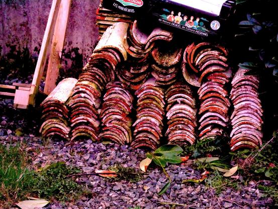 The image shows a stack of terracotta roof tiles arranged in a neat pile. They are placed on a bed of gravel, with some greenery around, including grass and a few leaves. There is also a wooden structure partially visible on the left side.