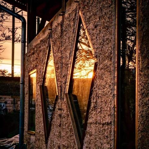 The image shows a wall with diamond-shaped windows during a sunset. The windows reflect the warm light and scenery outside, creating an aesthetically pleasing view. The wall appears to have a textured surface, possibly made from stone or a rough plaster material.