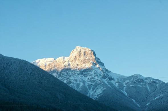 A late-sun, illuminated, jagged snow-capped peak from the Albertan Canadian rockies, surrounded by slightly shorter mountains which are shrouded in shadow.