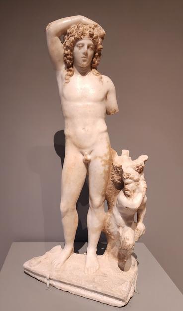 Marble statue of standing nude man with thick wavy hair. His left arm is missing and his right arm is lifted with his forearm resting on the top of his head. Beside him is a small marble Pan statue.