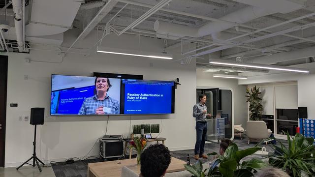 A person presenting using a computer. I. Their back two tv monitors one capturing their face and the other the presentation (with a blue background)