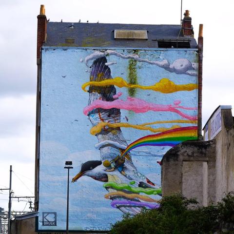 This image shows a large mural painted on the side of a building. The artwork features a stylized and colorful depiction of a bird with vibrant paint streaks trailing behind it. The background includes a cloudy sky, and colorful ribbons seem to intertwine with the bird in an abstract design. The mural is signed with the name "Brusk."