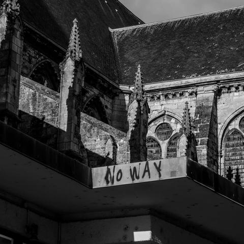 The image shows a black and white photograph of an old Gothic-style building, likely a church, with arched stained glass windows and ornate architectural details. In the foreground, there's a modern structure with graffiti that says "NO WAY." The combination creates a contrast between the historic and contemporary elements.