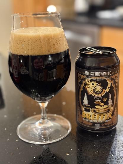 A tall snifter glass of dark beer with a thick tan bubbly head. The black can has a dark label with tan pancakes and a curious creature face